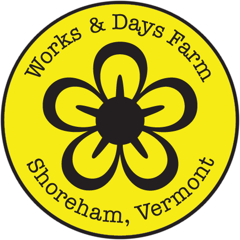 The logo of Works & Days Farm. A black-and-yellow flower with five petals inside a yellow circle with a black border. 'Works & Days Farm' is written at the top, and 'Shoreham, Vermont' is written at the bottom, both in black text.